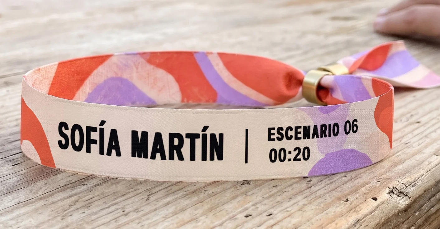 Detalle de la pulsera de boda festival: nombre del invitado, número de mesa y hora sobre ondas coral y lila